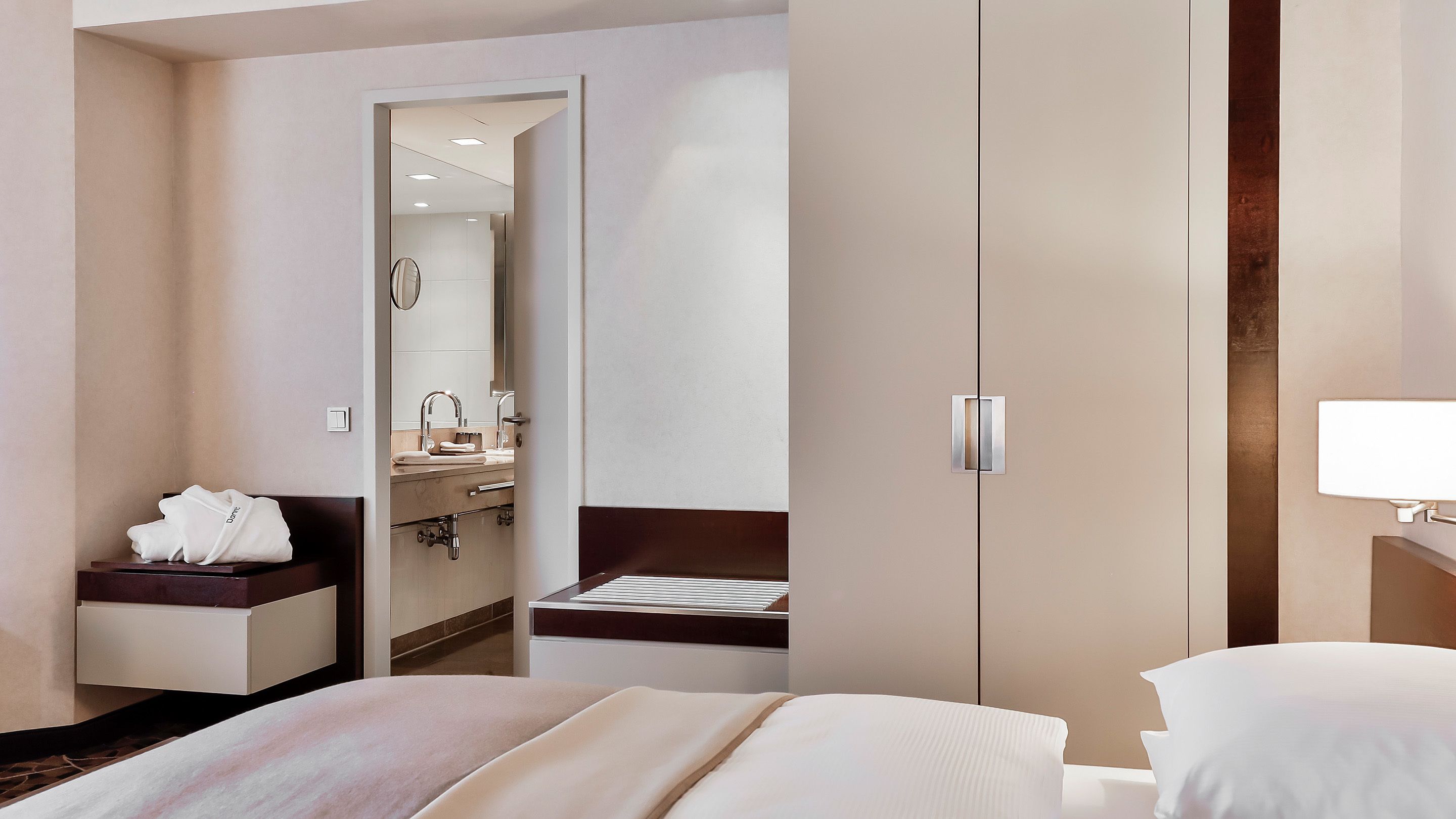 A hotel room with a bed, a bedside table with a lamp, a bathroom with a sink and mirror, and a wardrobe.
