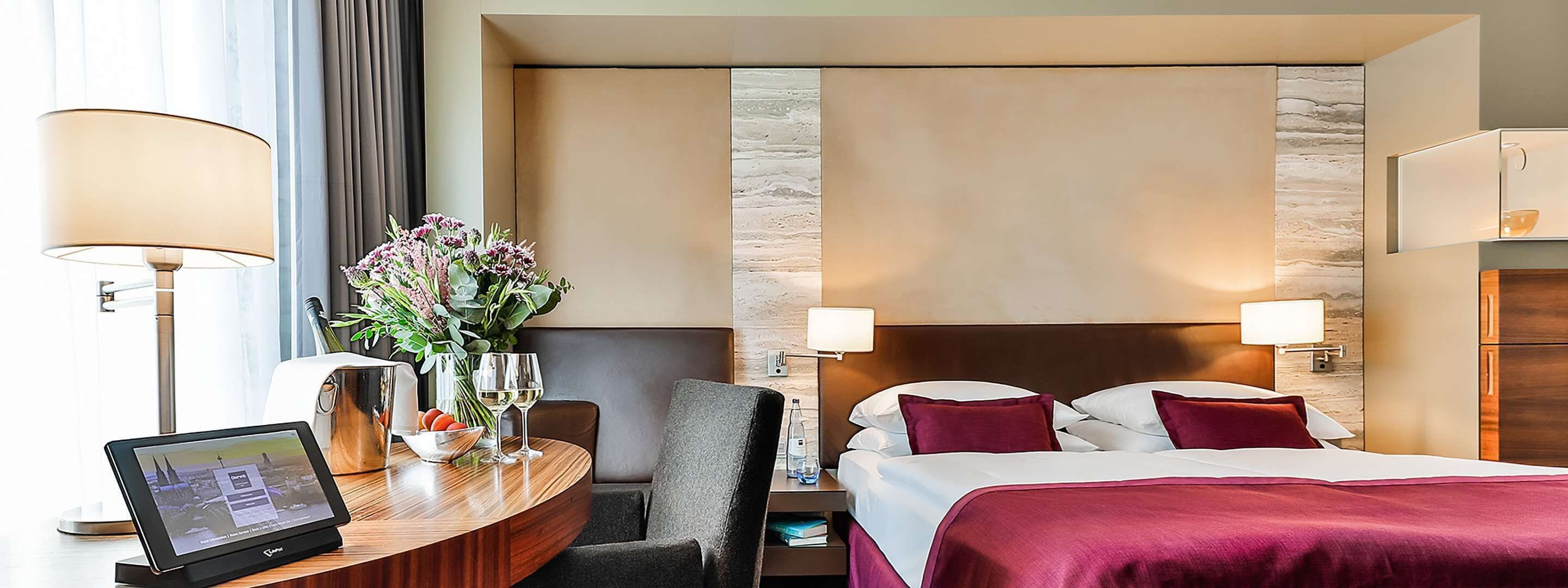 Habitación de hotel con cama, mesitas de noche, escritorio con computadora portátil, un florero con flores y vistas a la ciudad.