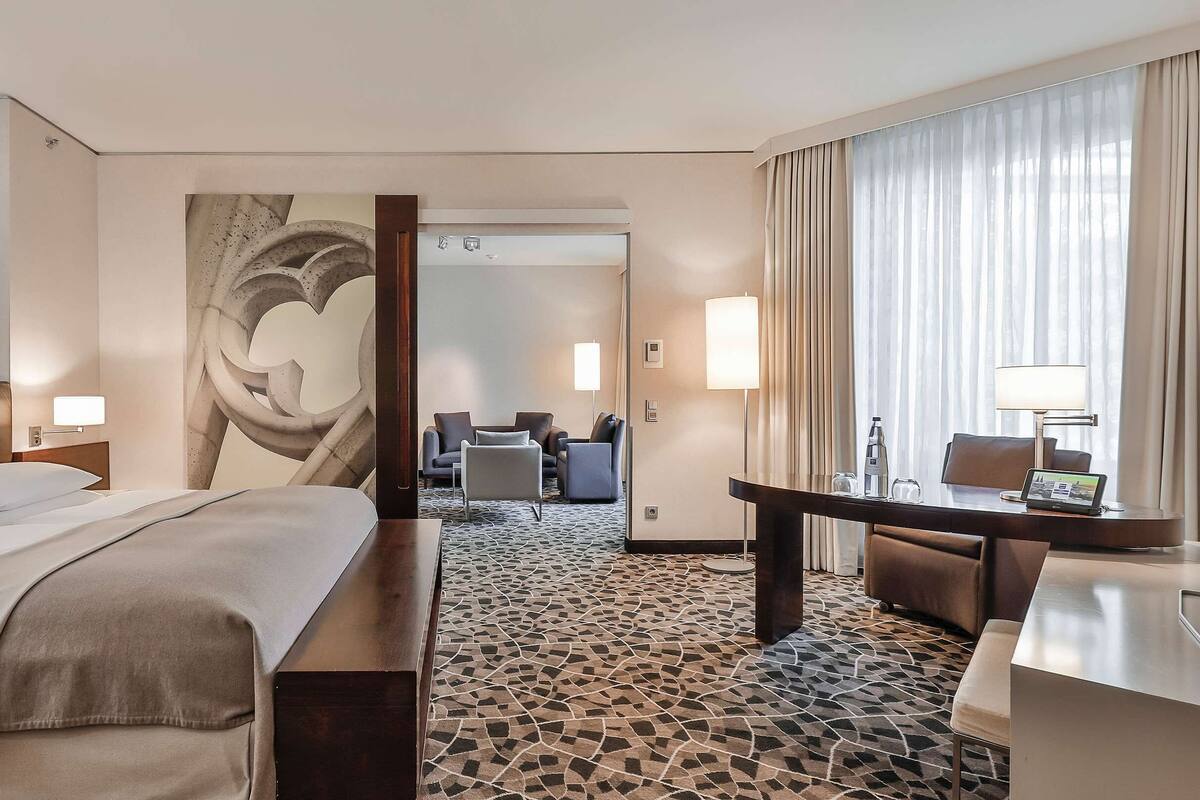 A hotel room with a bed, a desk, a chair, a sofa, and a large window with curtains.