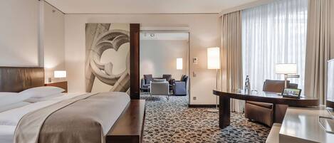 A hotel room with a bed, a desk, a chair, a sofa, and a large window with curtains.