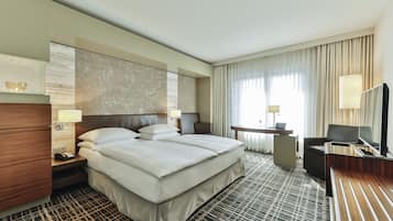 A hotel room with a large bed, bedside tables, a desk with a chair, a TV, and a window with curtains.