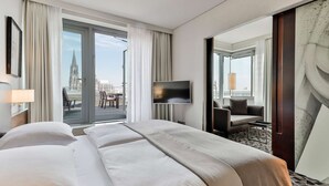 A hotel room with a large bed, a TV, a seating area with a chair and table, and a view of a cityscape through the windows. - Dorint Hotel am Heumarkt Köln (Cologne)