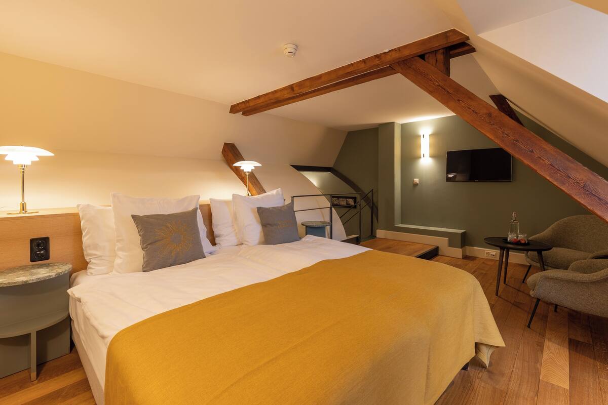 A bedroom with a bed, bedside table, lamp, chair, and a TV on the wall.