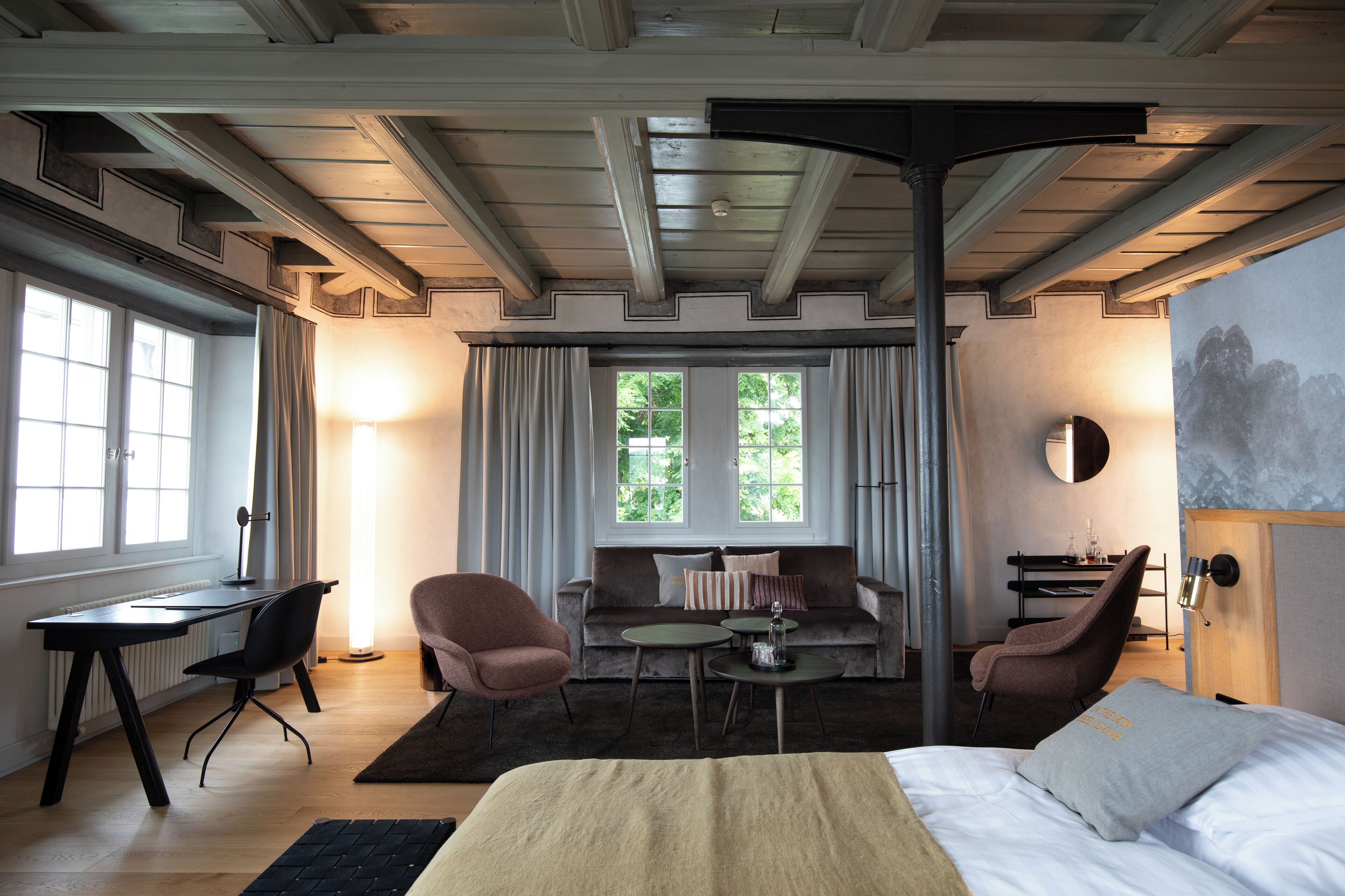 Ein Hotelzimmer mit einem Bett, einem Schreibtisch, zwei Stühlen, einem Sofa und einem großen Fenster mit Vorhängen.