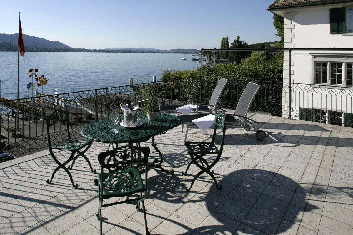 A terrace with outdoor seating, a view of a lake, and a building with a tiled roof.