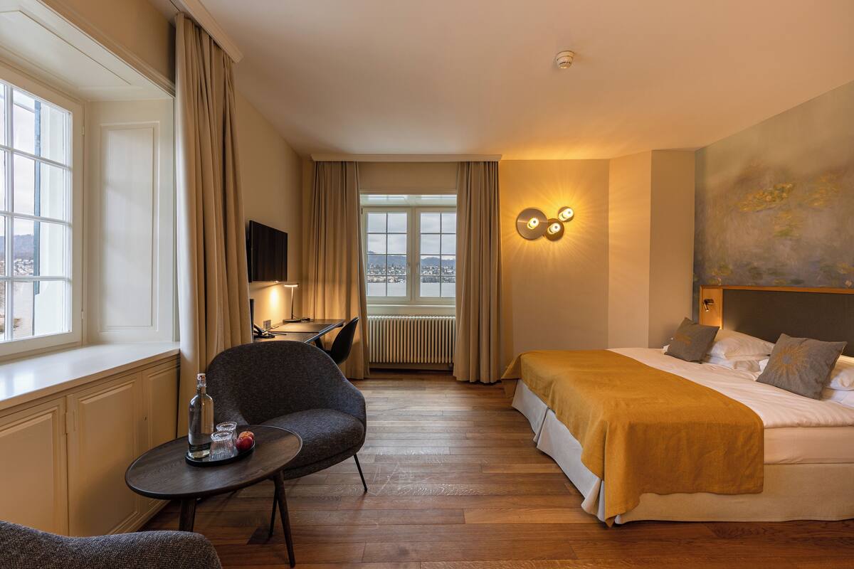 A hotel room with a bed, a desk with a chair, a small table with a bottle and fruit, and a window with curtains.
