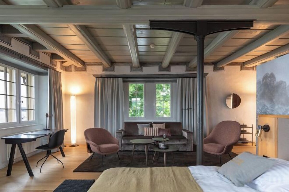 Una habitación con cama, dos sillones, un escritorio y un sofá. Cuenta con ventanas amplias y vistas al exterior con zonas verdes.