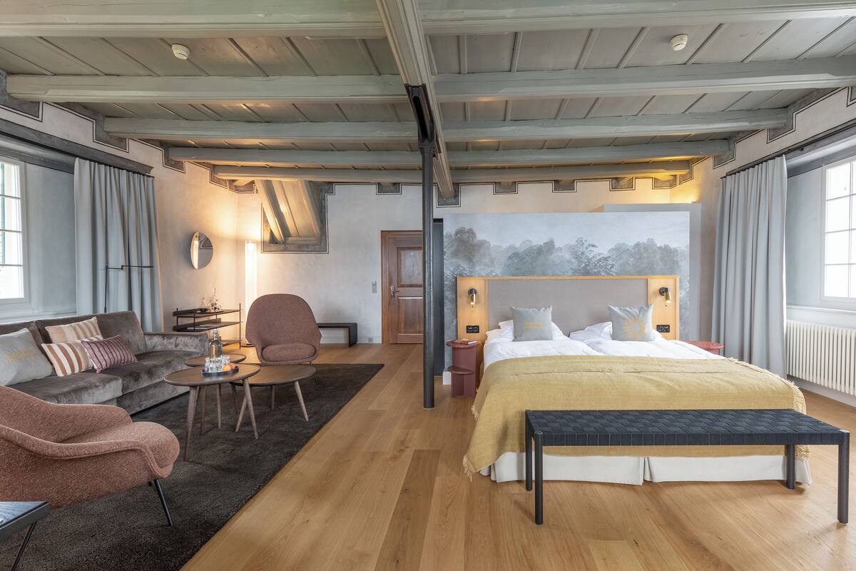 A modern hotel room with a large bed, a sofa, a chair, a small table, and a bench.