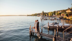 A couple standing on a wooden dock extending into a calm body of water, with boats docked nearby and a residential area in the background. - Seehotel Sonne (Kuesnacht)