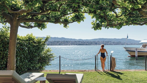 A woman in a swimsuit walking along a dock by a lake with lounge chairs and a boat named "TLIBERG" in the background. - Seehotel Sonne (Kuesnacht)