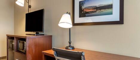 A hotel room with a wooden desk, a television, a lamp, a framed picture of a bridge, and a minibar.