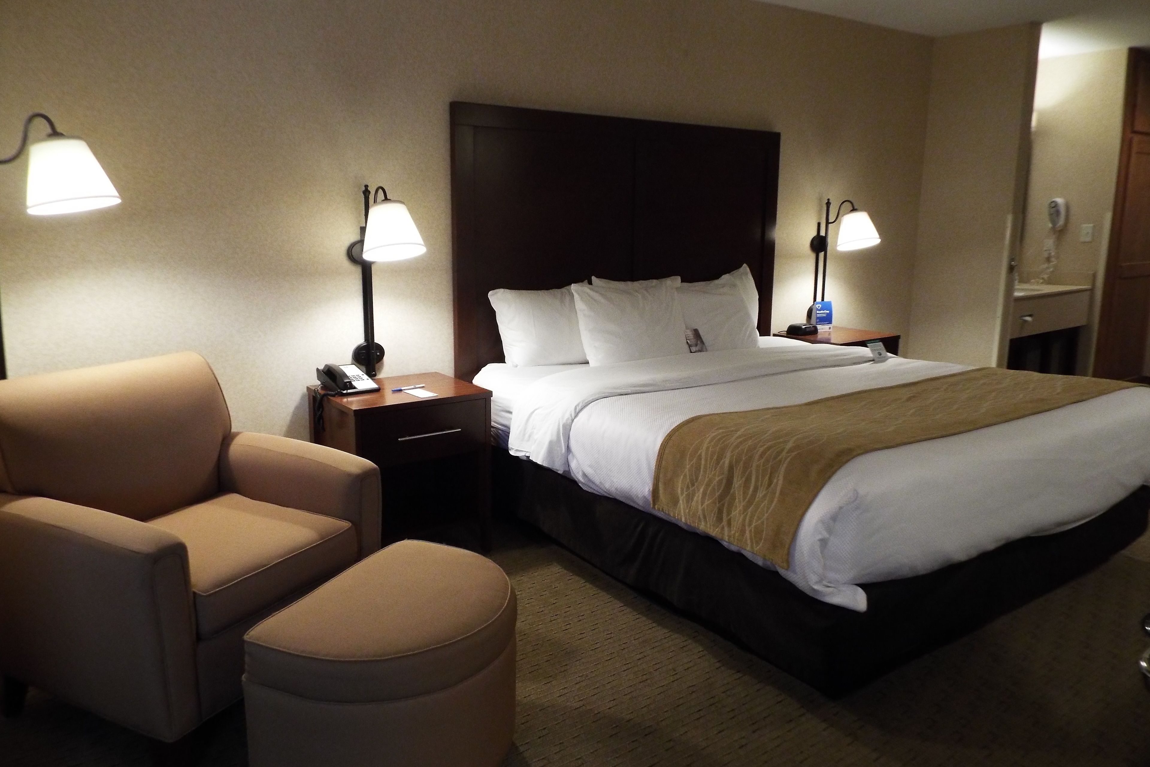 A hotel room with a large bed, two bedside lamps, a chair, and a small ottoman.