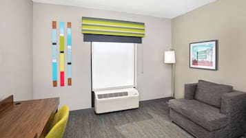 A hotel room with a wooden desk, a gray sofa, a large window with a striped blind, a standing lamp, and a framed picture on the wall.