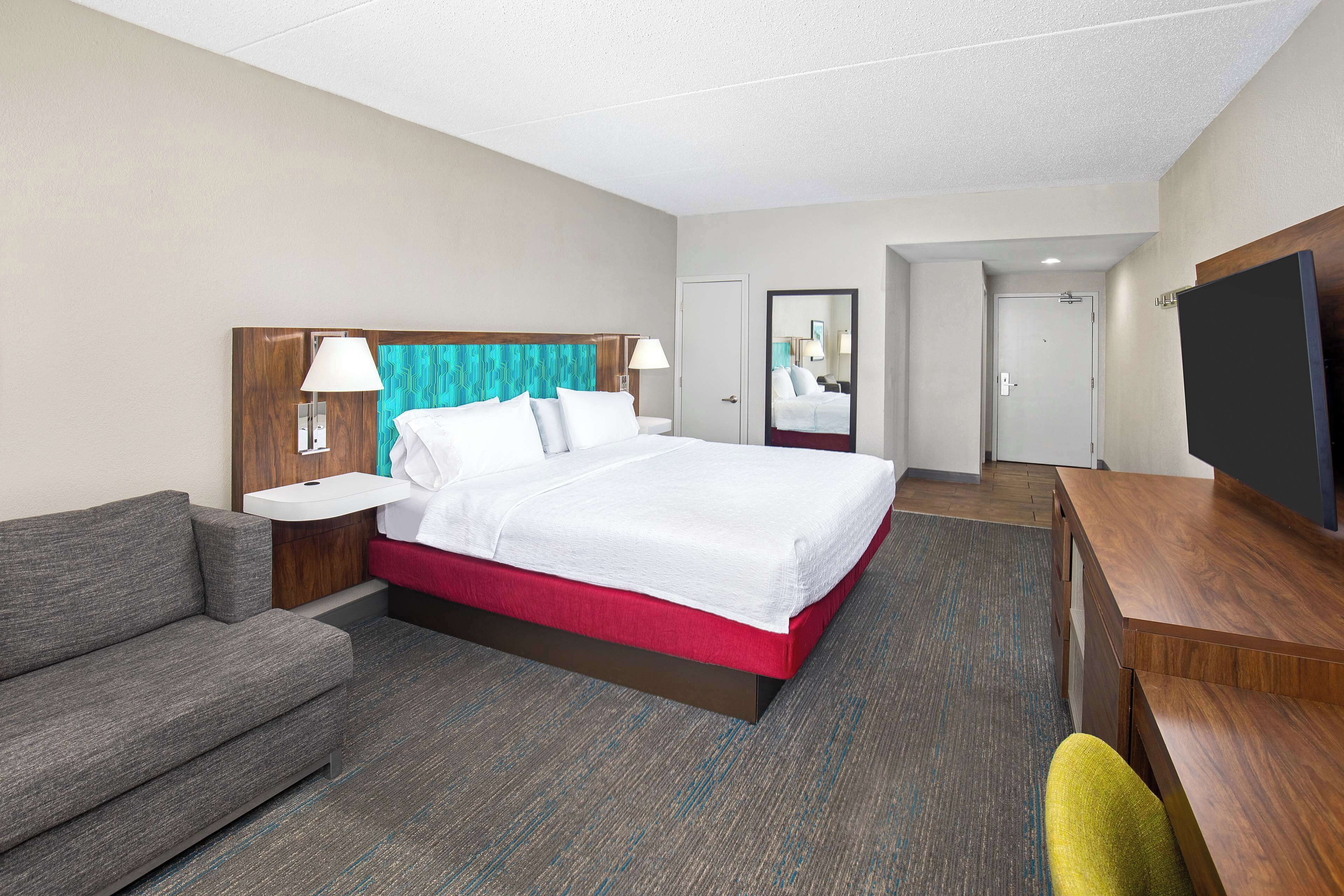 A hotel room with a bed, a sofa, a desk, a TV, and a bathroom visible through an open door.