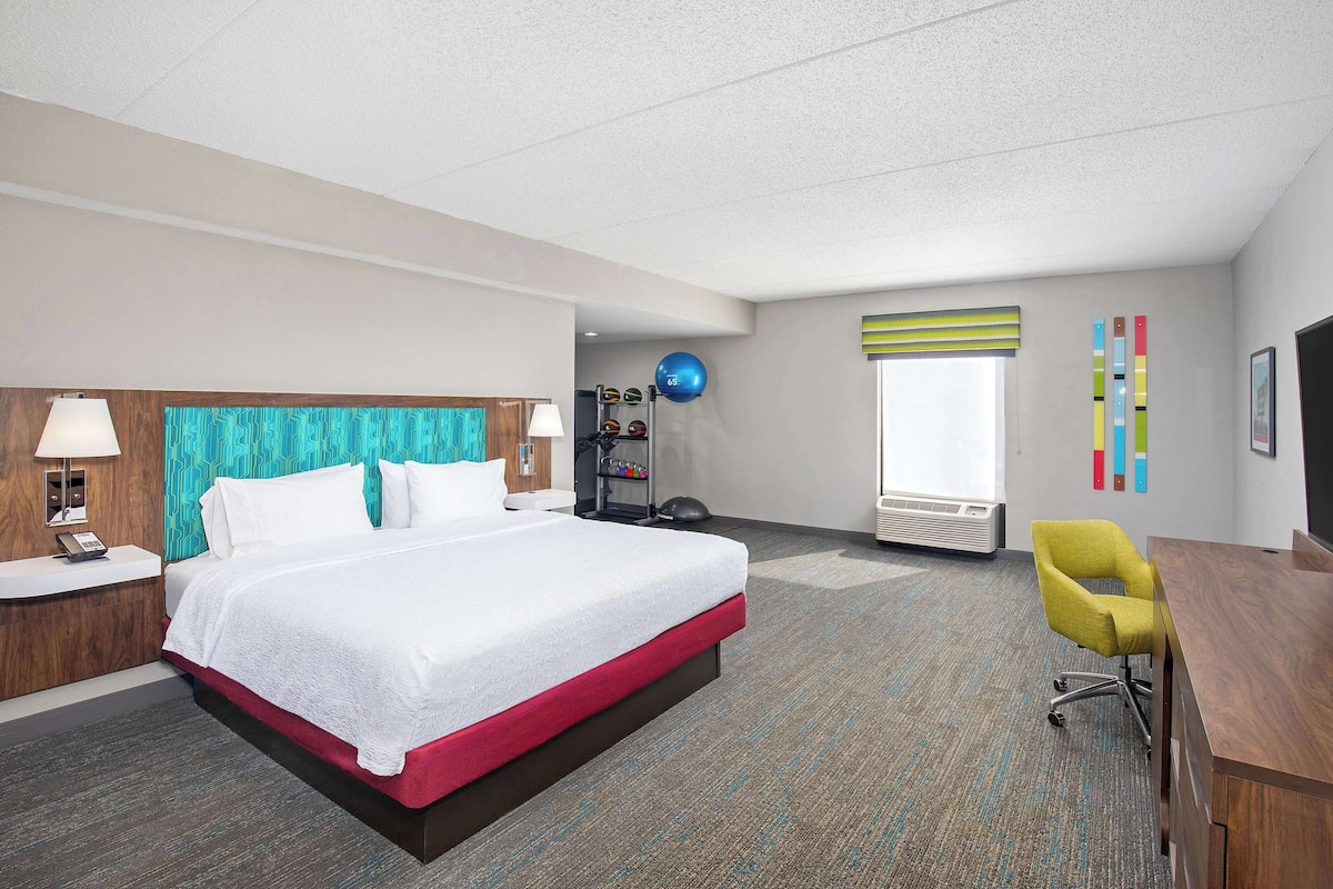 Quarto de hotel com uma cama grande, uma escrivaninha, uma cadeira, um abajur, uma televisão, uma janela com persianas, uma estante com artigos esportivos e uma parede com decoração colorida.