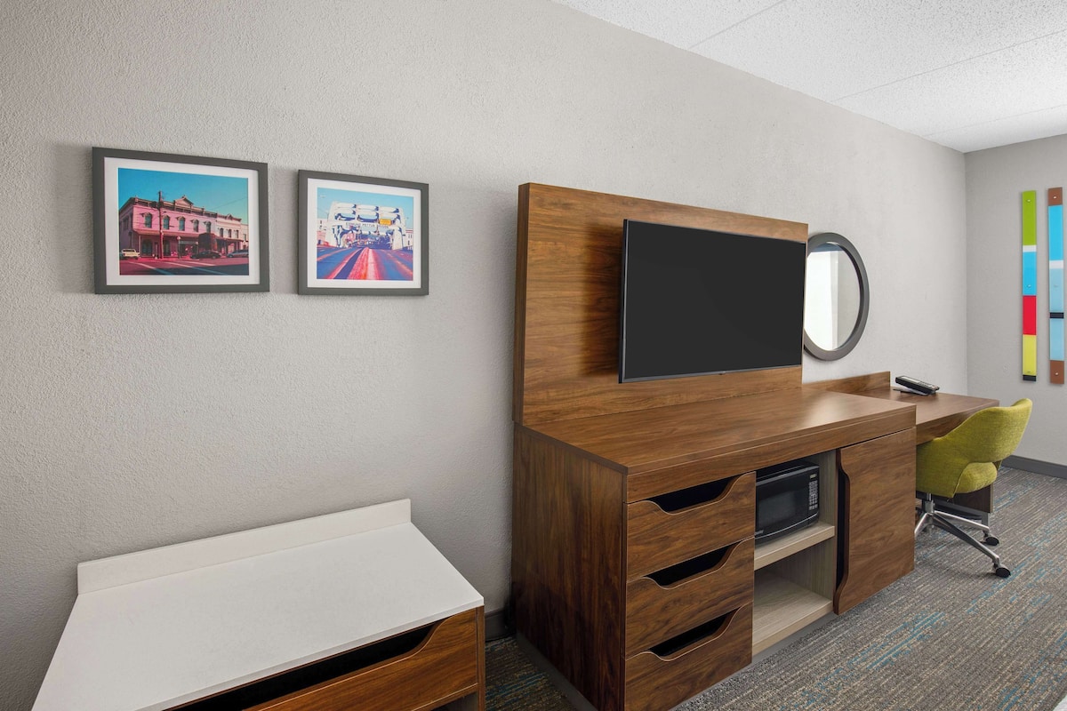 Quarto de hotel com escrivaninha de madeira, televisão de tela plana, mesa pequena branca e duas quadros na parede.