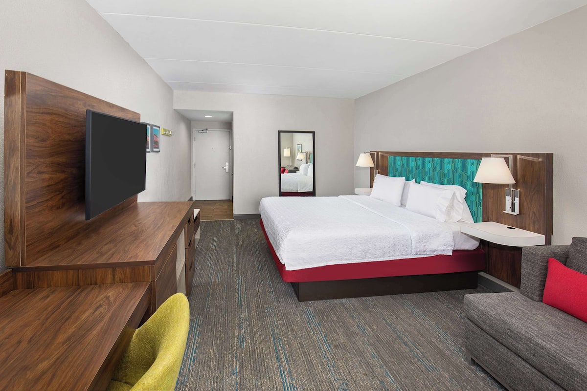 Quarto de hotel com cama, escrivaninha, televisão, espelho e sofá.