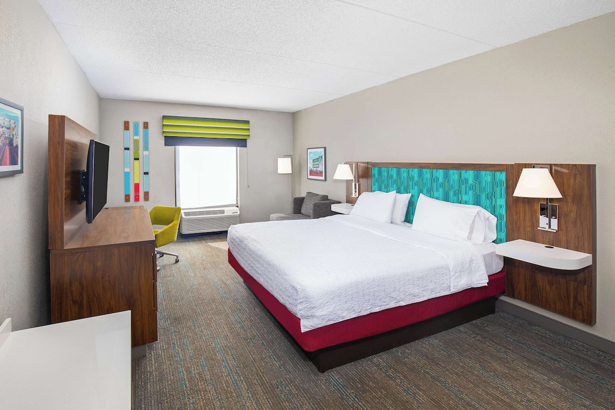 Quarto de hotel com uma cama grande, uma escrivaninha, uma cadeira, uma televisão e uma janela com persianas.