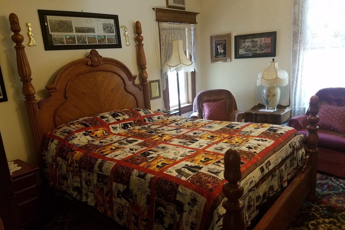 A bedroom with a wooden four-poster bed, a quilt with various images, a chair, a nightstand, and a lamp.