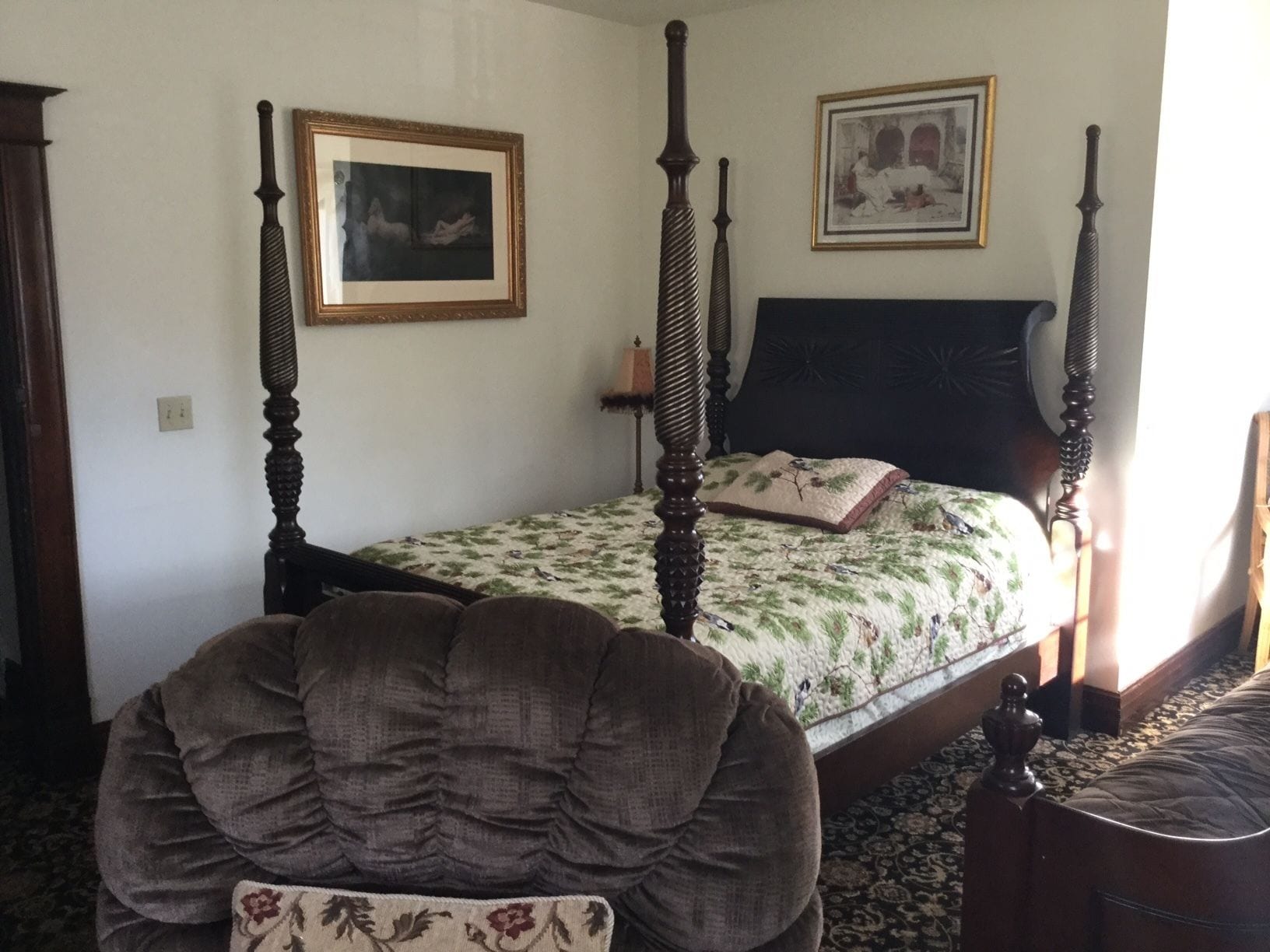 A four-poster bed with a floral bedspread and a single pillow, a framed picture above the bed, and a chair in the foreground.