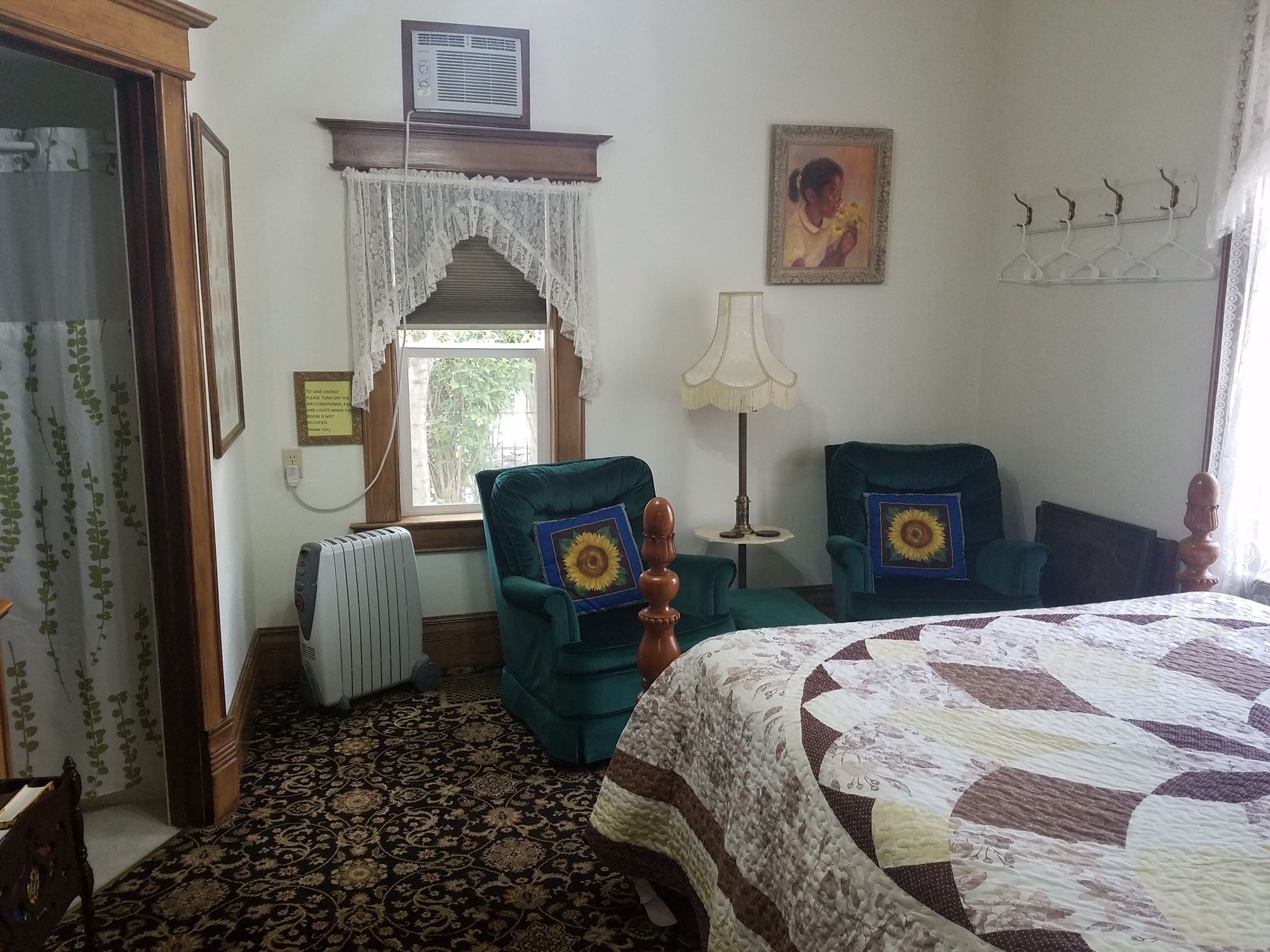 A hotel room with a large bed, two green armchairs, a radiator, a window with lace curtains, a wall-mounted air conditioner, and a framed picture.