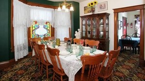 A dining room with a large wooden table set for six, surrounded by six wooden chairs. There is a stained glass window with a floral design behind the table, and a wooden cabinet with glass doors to the right. - Spurs n Lace Bed & Breakfast Inn (Colorado Springs)