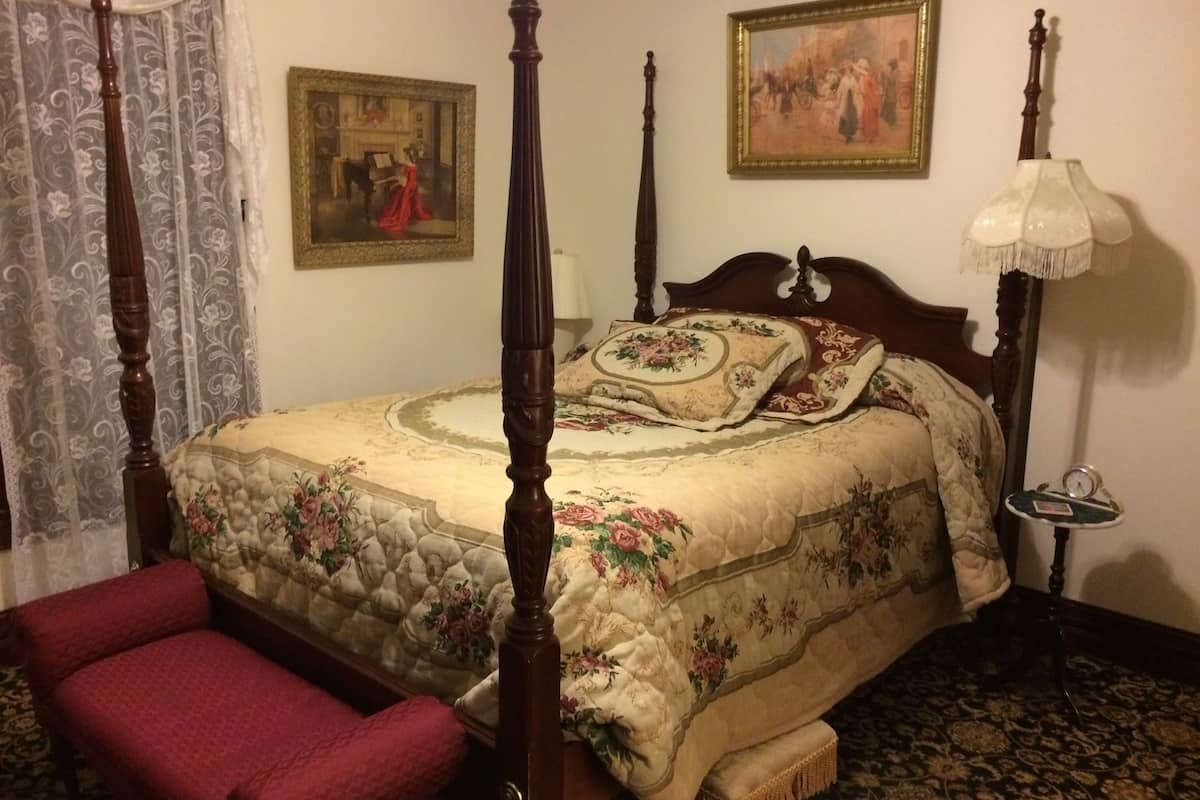 A four-poster bed with floral bedding, a red upholstered bench, and a painting above the bed.