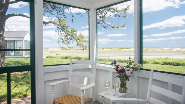 A white porch with a bench and a table with a vase of flowers and two glasses of wine. - Dunes on the Waterfront (Ogunquit)