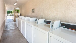 A laundry area with multiple white washing machines and dryers, a sign indicating the availability of laundry soap and suds, and a concrete floor.