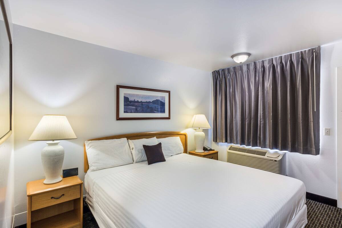 A hotel room with a large bed, two bedside tables with lamps, a framed picture on the wall, a window with curtains, and a ceiling light.