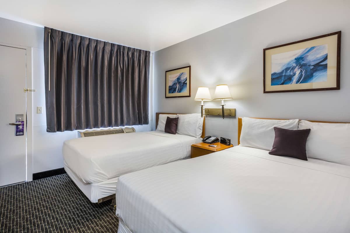 A hotel room with two beds, a nightstand with a telephone, two wall-mounted lamps, and a large window with curtains.