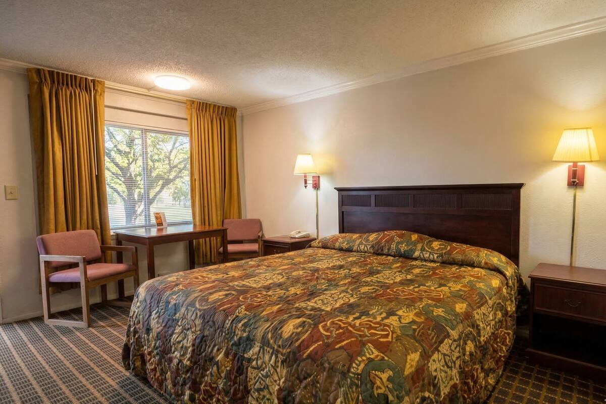 Ein Hotelzimmer mit einem großen Bett, zwei Nachttischen, einem Schreibtisch mit Stuhl und einem fenster mit Vorhängen.