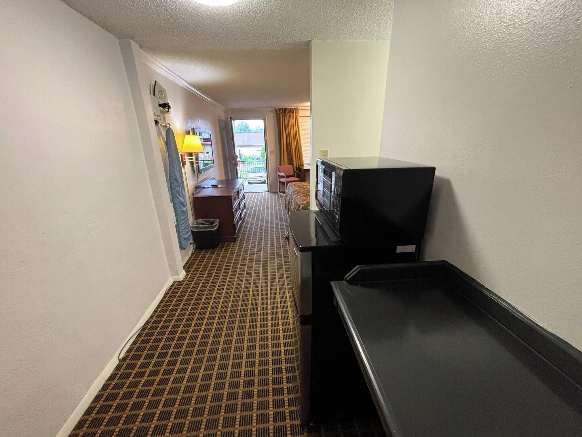Pasillo de hotel con alfombra estampada, televisor sobre un soporte, un cubo de basura y un escritorio con lámpara.