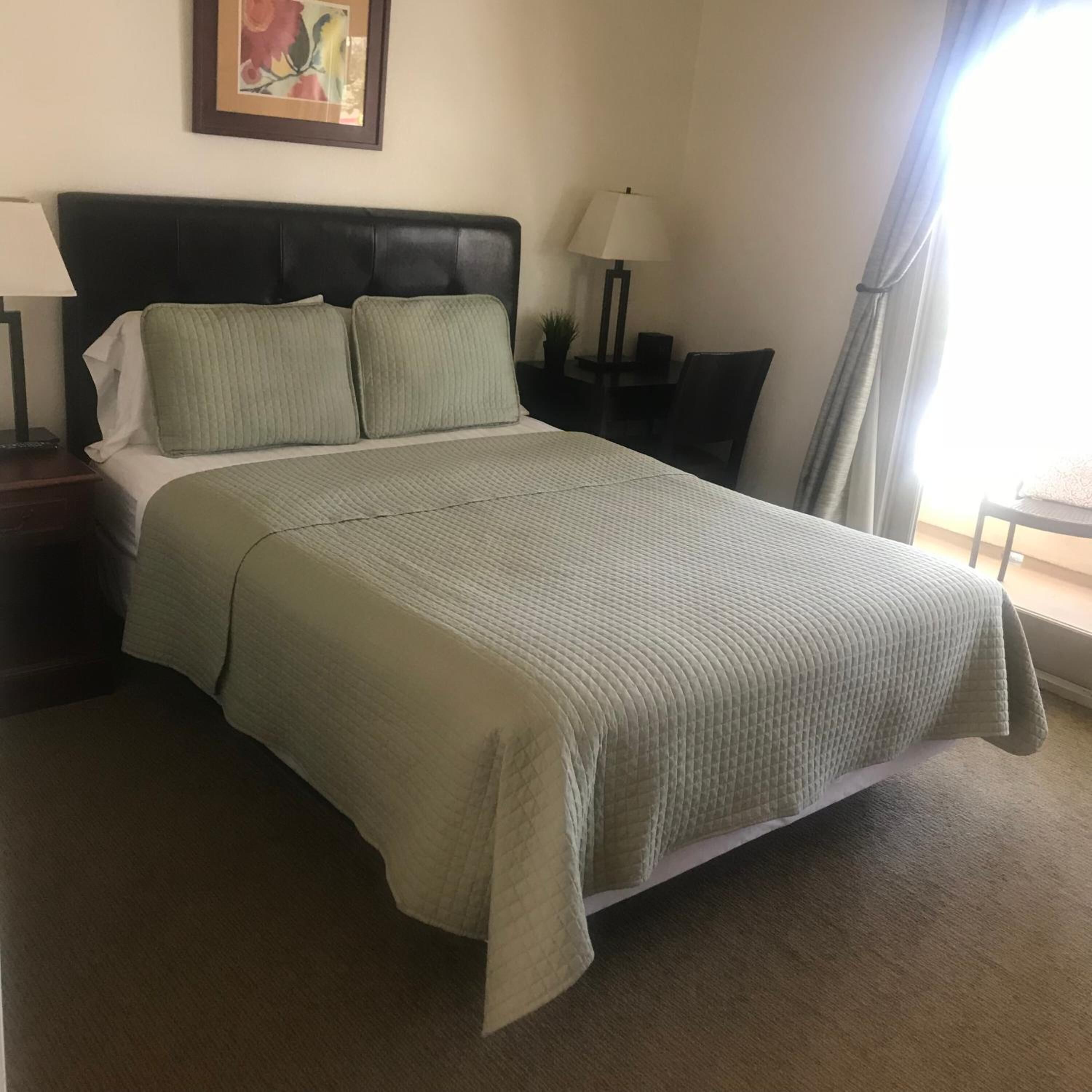 Una cama bien hecha con colcha acolchada y almohadas, una mesita de noche con lámpara y un cuadro en la pared.