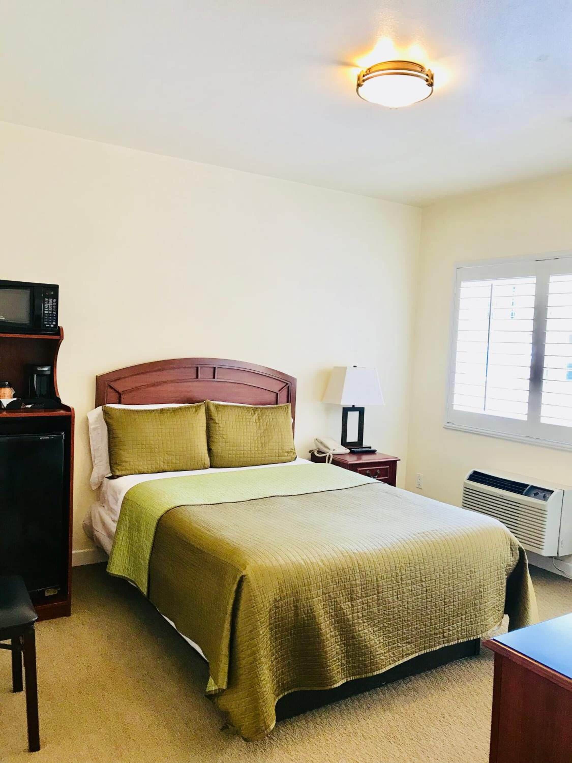 A hotel room with a bed, a bedside table with a lamp, a desk with a microwave, and a window with blinds.