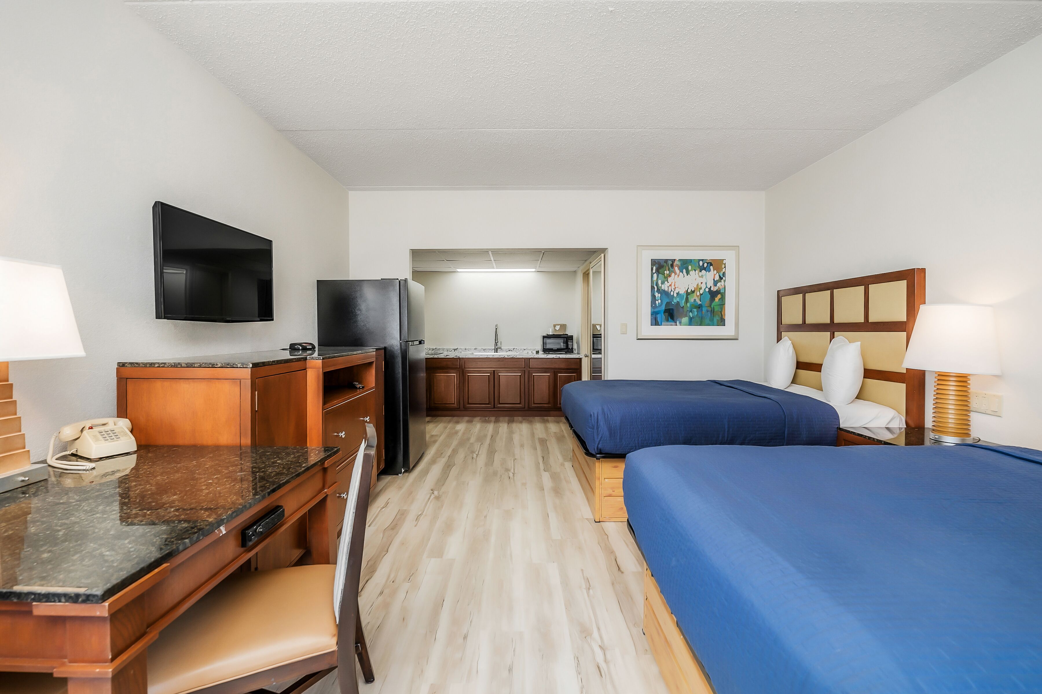 Una habitación de hotel compacta con kitchenette, un escritorio con teléfono, una cama con cubrecama azul y un cuadro en la pared.