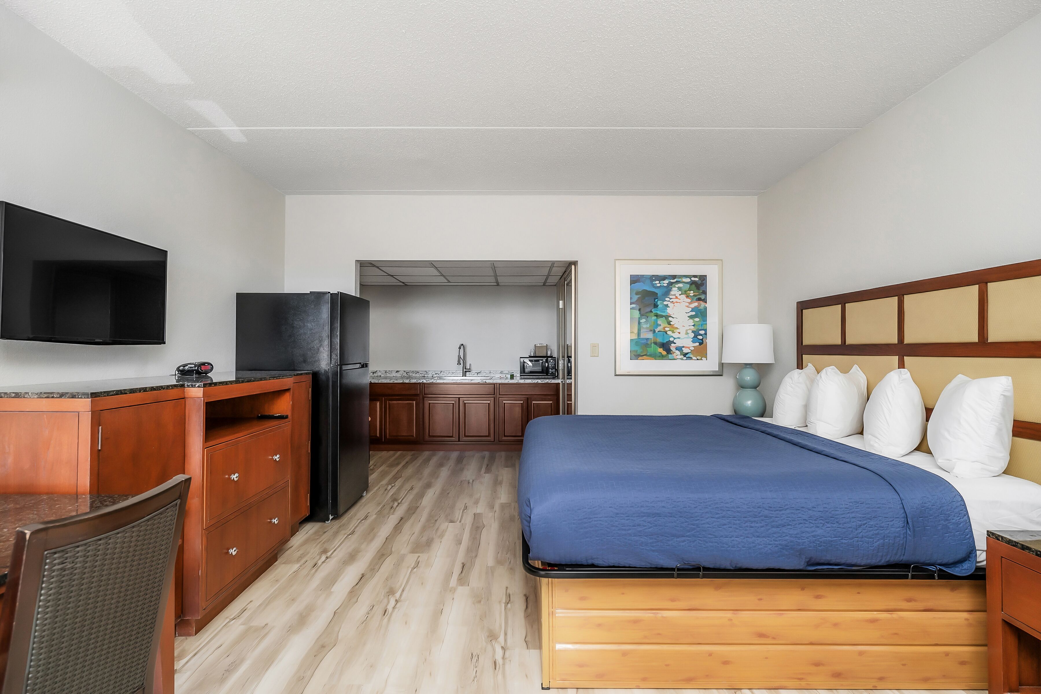 A hotel room with a bed, a desk, a TV, a refrigerator, a sink, and a painting on the wall.