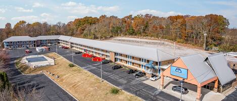 A motel with a large parking lot, a covered outdoor seating area, and a view of autumn trees in the background.