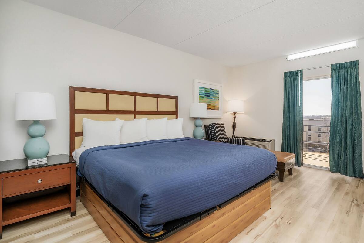 Une chambre d’hôtel avec un grand lit, une table de chevet, une lampe, une chaise et une fenêtre avec des rideaux.
