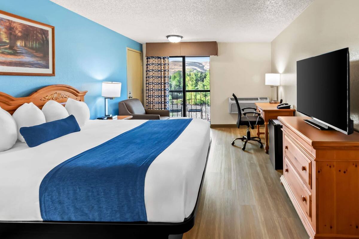 Une chambre d’hôtel avec un grand lit, un bureau avec un ordinateur, une télévision et un balcon donnant sur la verdure.