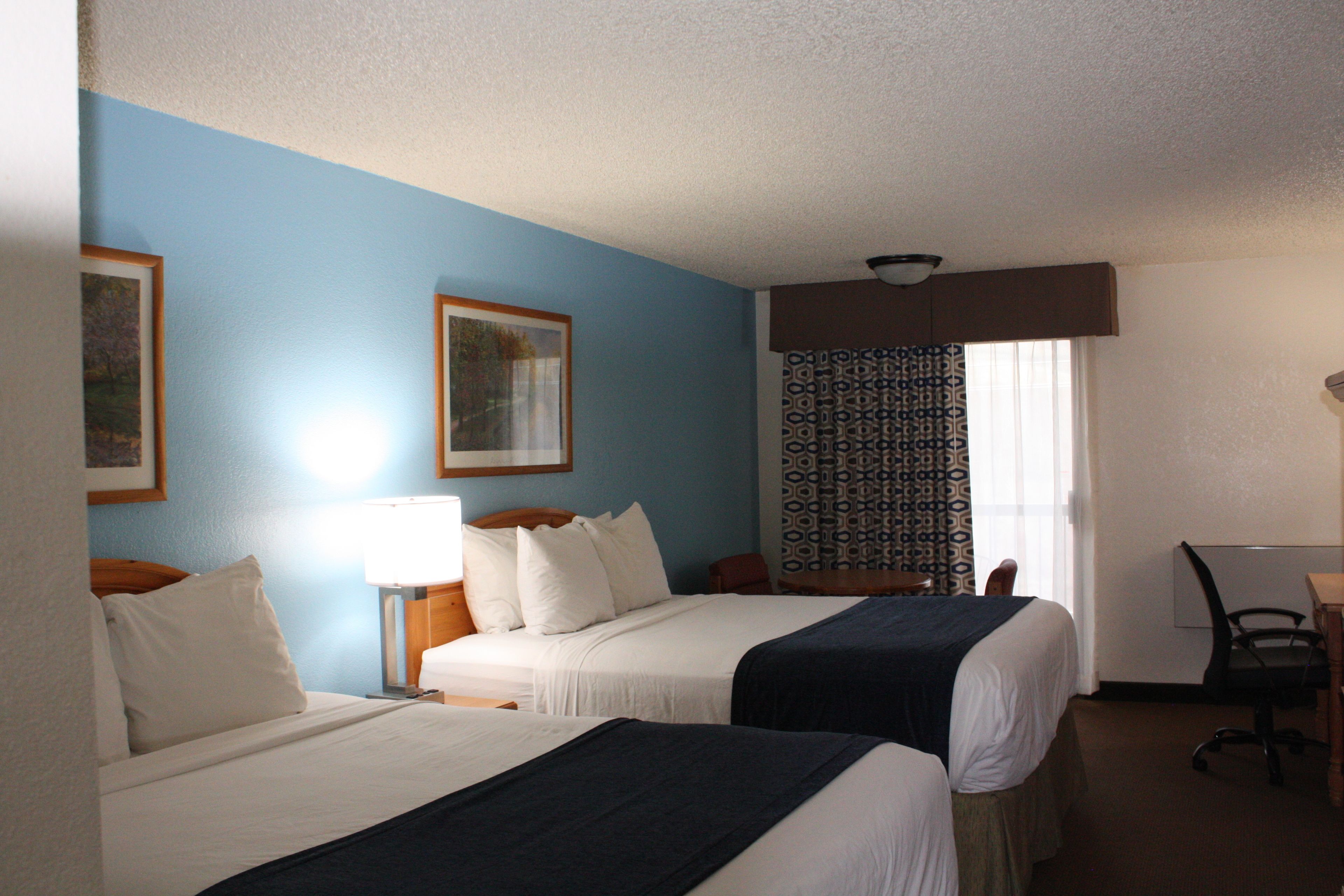 A hotel room with two beds, a desk, a chair, and a window with patterned curtains.