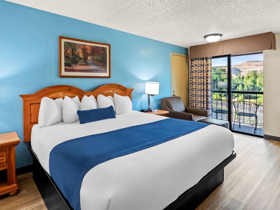 A hotel room with a large bed, a painting on the wall, a balcony with a view, and a chair.