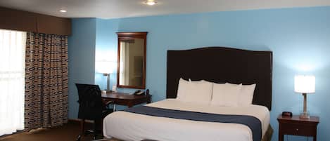 A hotel room with a large bed, a desk with a chair, a bedside table with a lamp, and a window with patterned curtains.