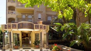 Entrada de un hotel con un arco decorativo, plantas en macetas y un jardín con plantas y flores tropicales.
