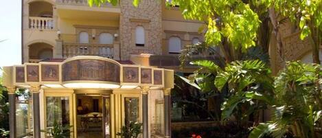 Entrada de un hotel con un arco decorativo, plantas en macetas y un jardín con plantas y flores tropicales.