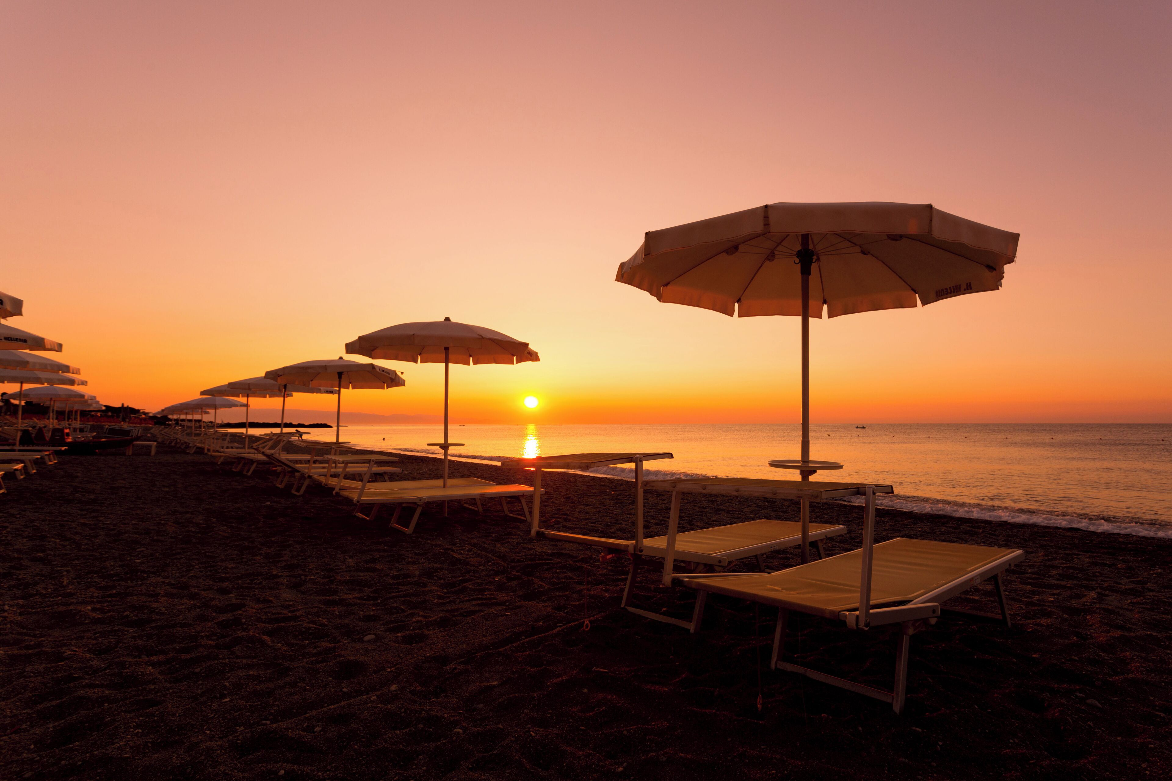 A beach with rows of sun loungers and umbrellas, a sunset in the background, and the ocean visible.