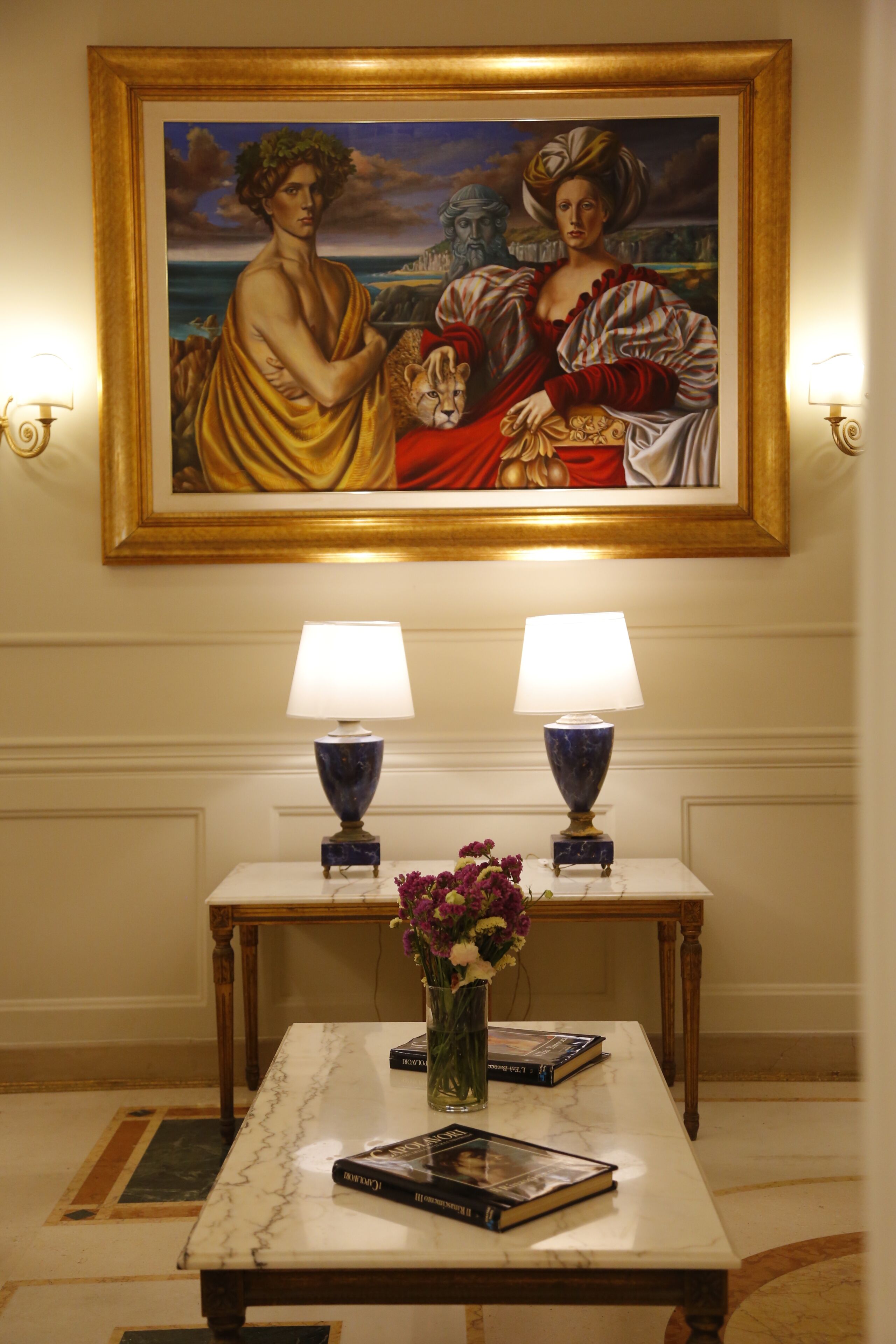 Una stanza con un tavolo in marmo, due lampade, un vaso con fiori e un quadro appeso al muro.