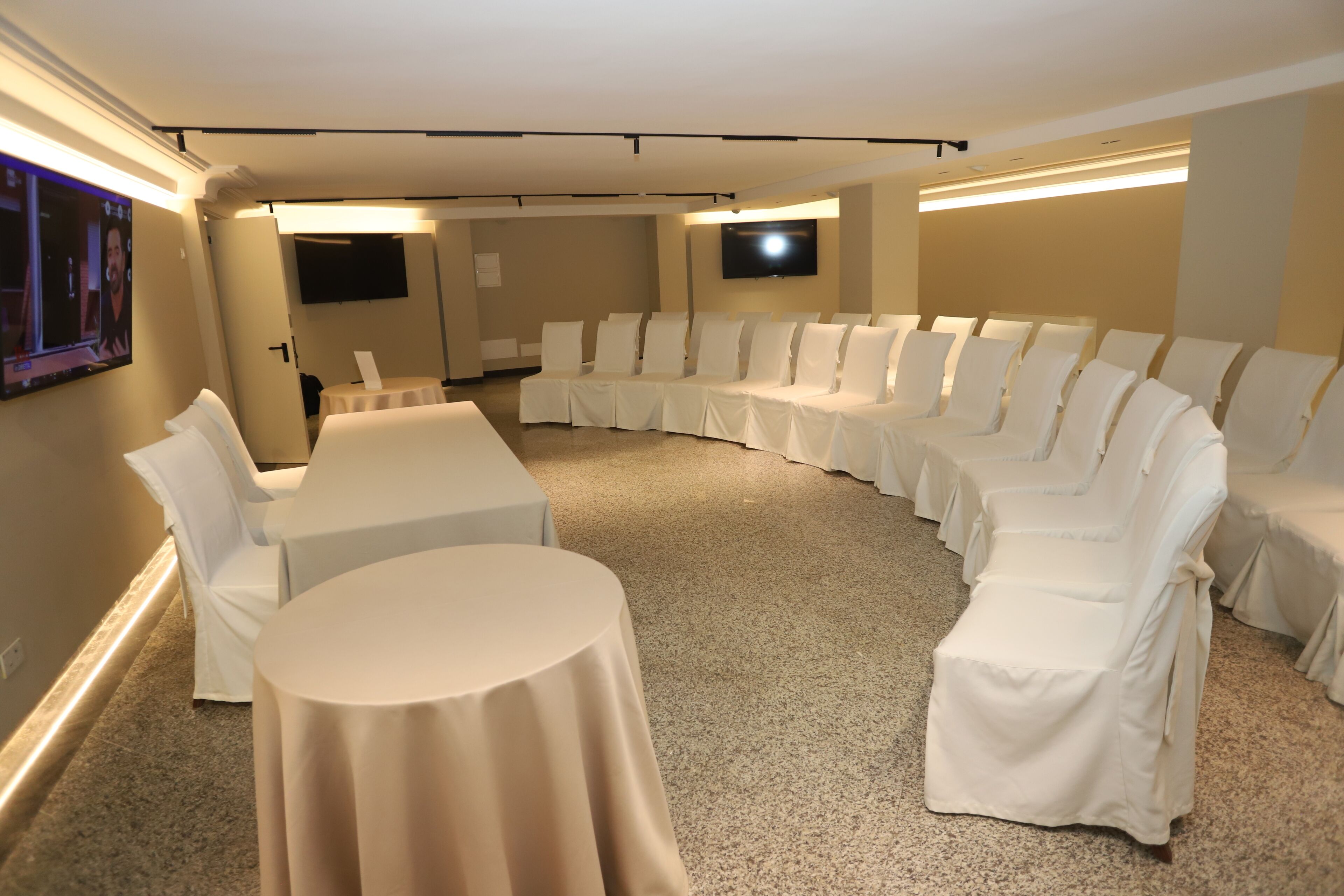 Un salón de conferencias con una mesa blanca curva y sillas, piso alfombrado y una pantalla plana montada en la pared.