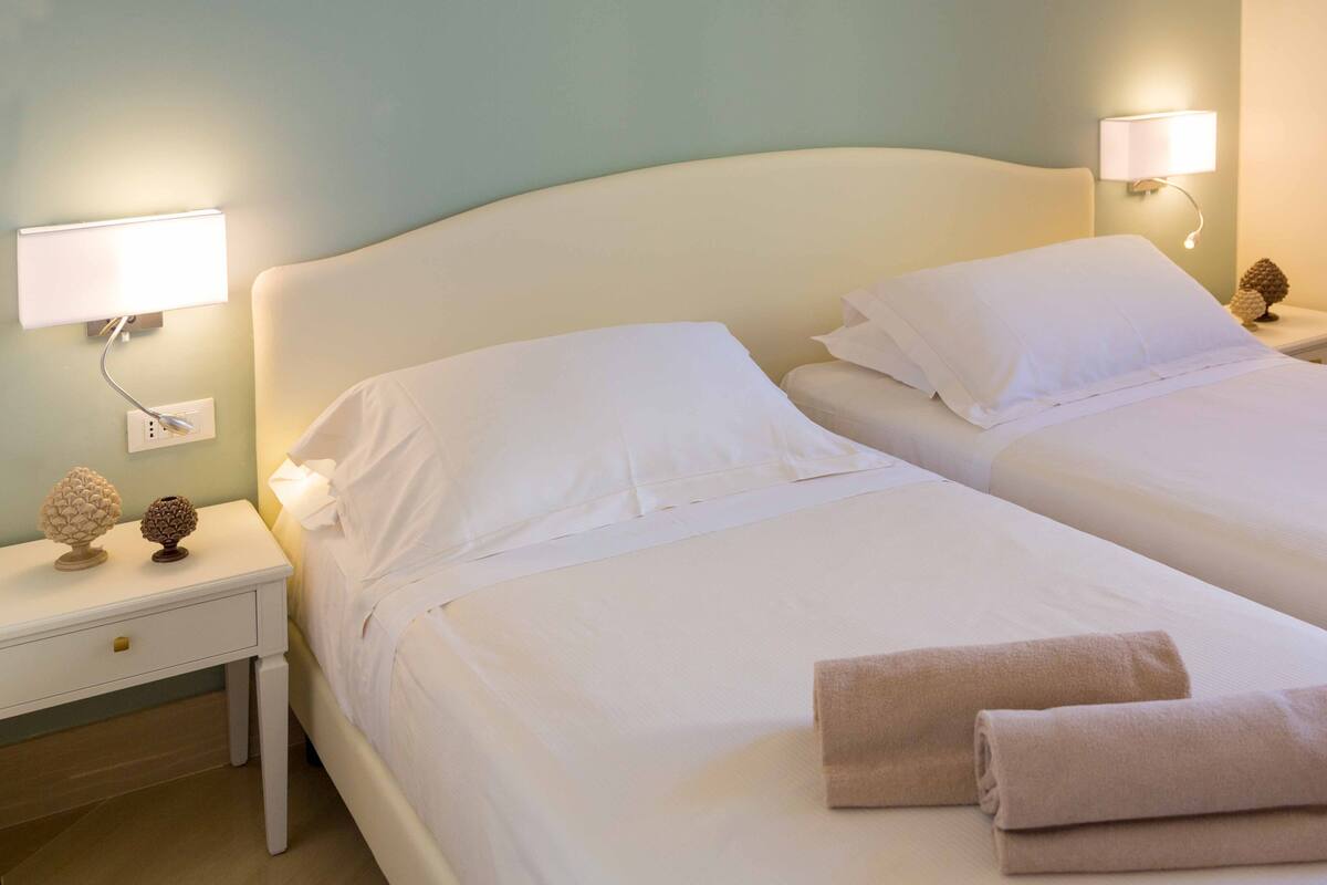 Een tweepersoonsbed met witte lakens en twee kussens, een nachtkastje met een lamp, en een wandlamp.