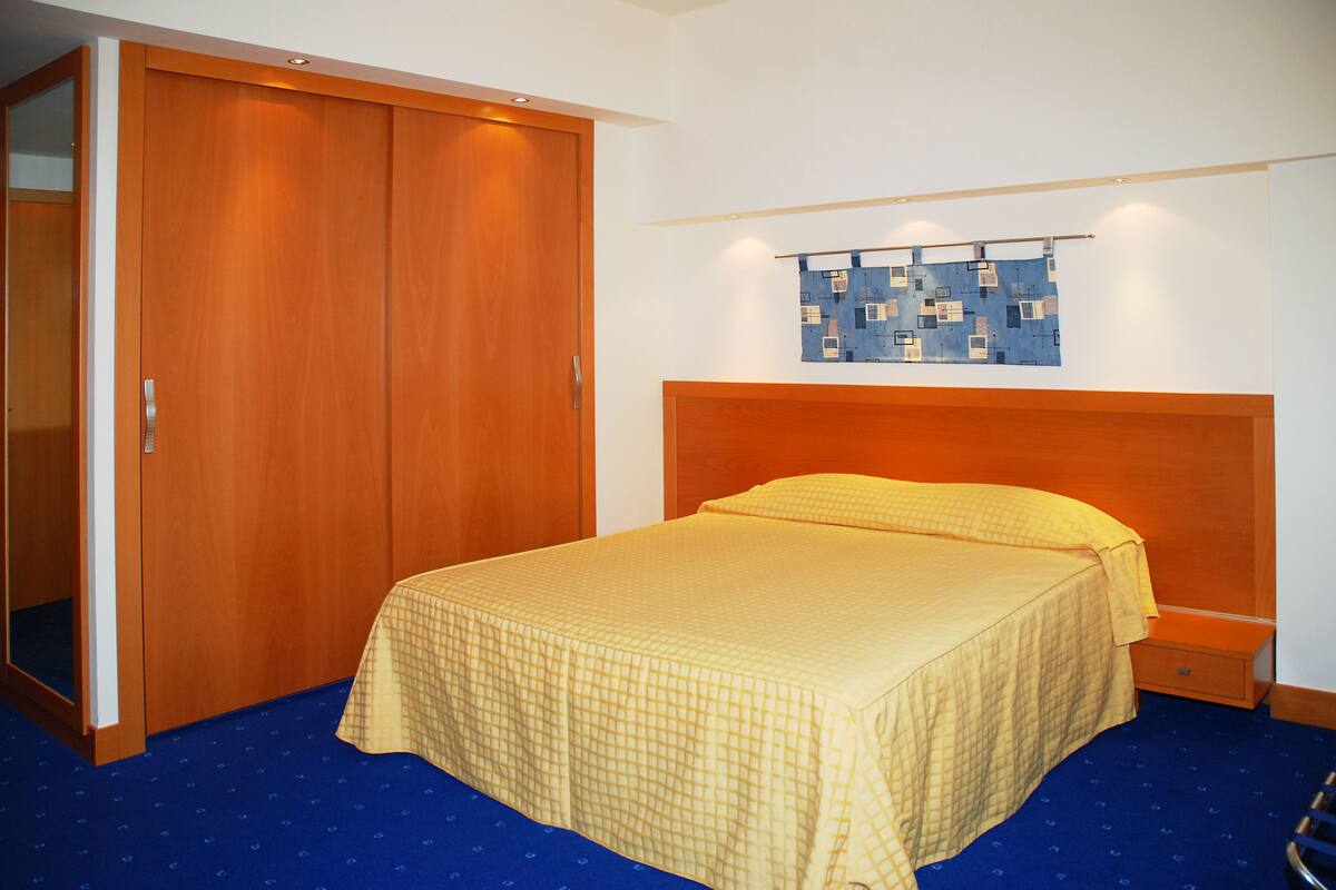 A hotel room with a large wardrobe, a bed with a yellow bedspread, and a blue carpet.